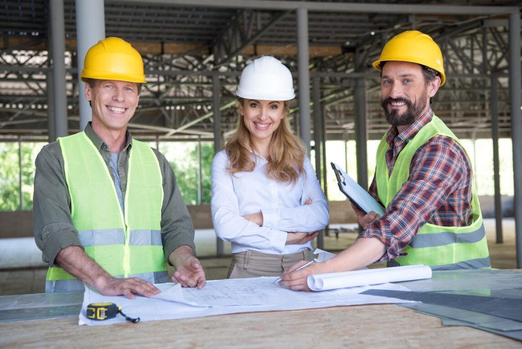 Three people wearing hard hats and safety vests stand in front of a large blueprint, engaged in a discussion