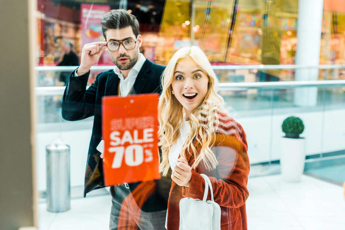 Inside a store, a man and woman are focused on a sale sign, indicating their interest in the promotional items available.