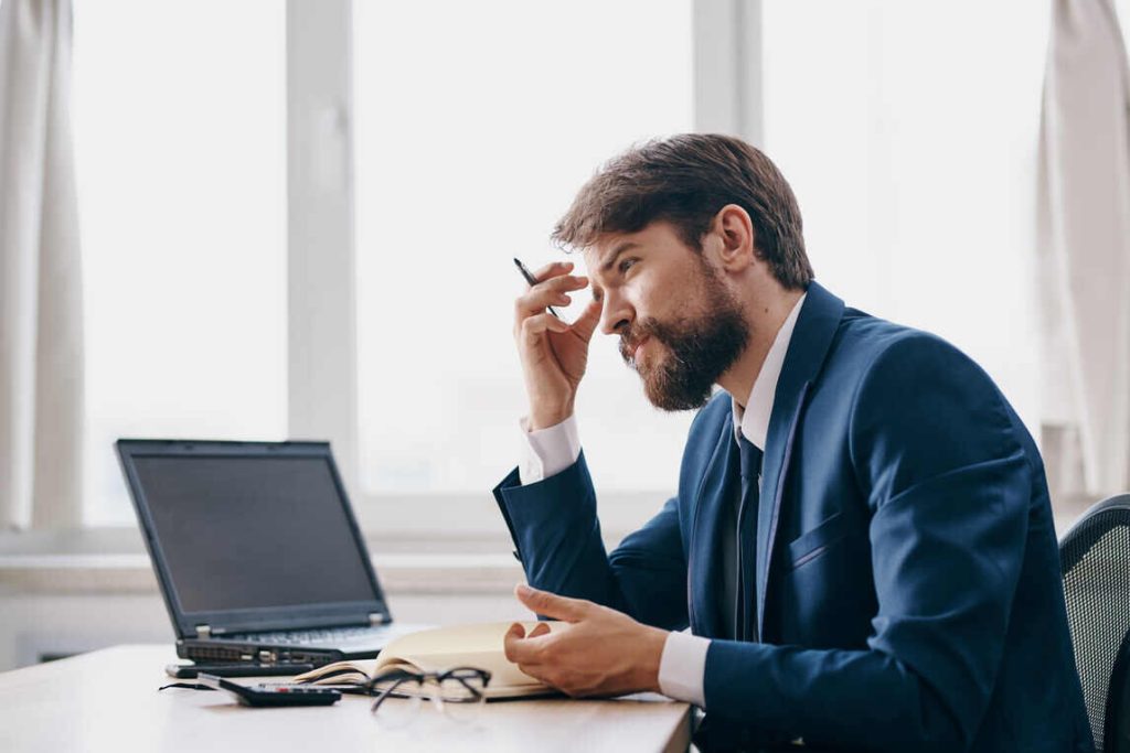 A man in a suit sits at a desk, working on his laptop with a focused expression.